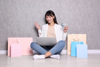 Internet shopping. Happy woman with credit card, laptop and colorful bags sitting near grey wall