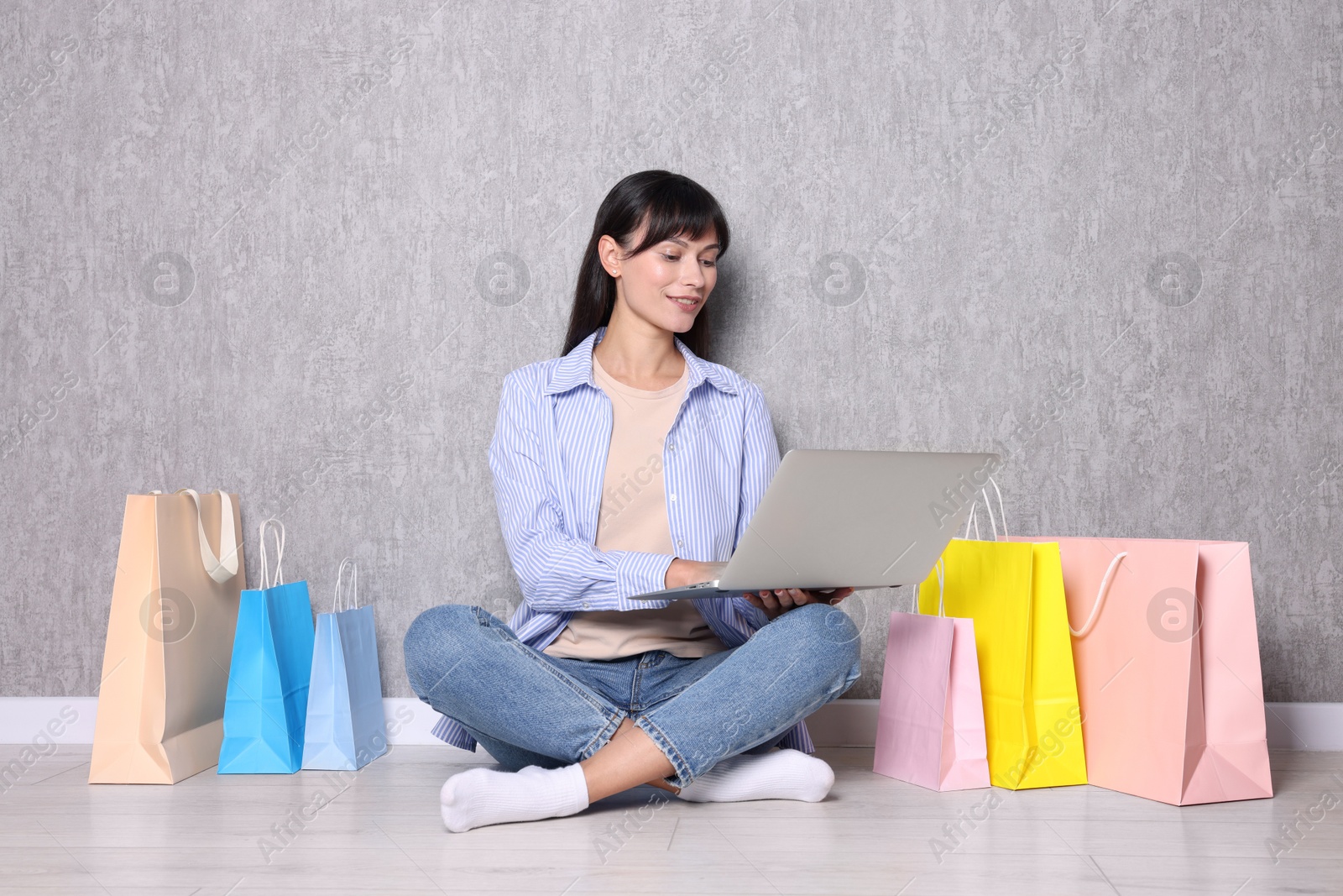 Photo of Internet shopping. Happy woman with laptop and colorful bags sitting near grey wall