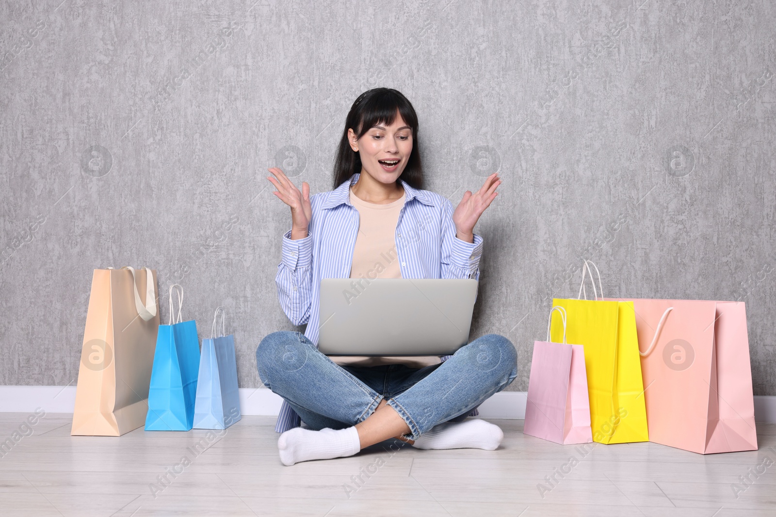 Photo of Internet shopping. Happy woman with laptop and colorful bags sitting near grey wall