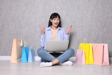 Internet shopping. Happy woman with laptop and colorful bags sitting near grey wall