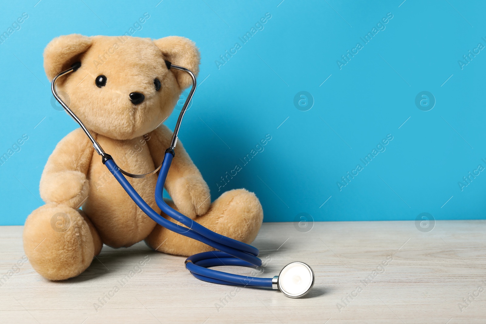 Photo of Pediatrics concept. Teddy bear with stethoscope on wooden table against light blue background, space for text