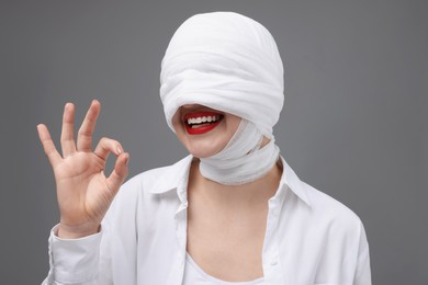 Woman with face wrapped in medical bandage after plastic surgery operation showing ok gesture on grey background