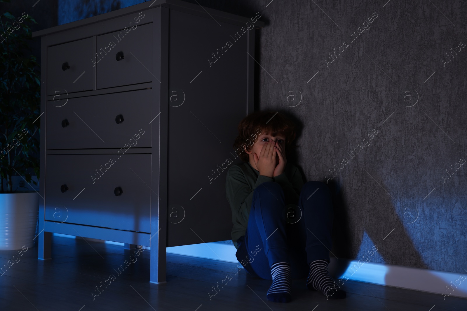 Photo of Scared boy hiding behind chest of drawers at night