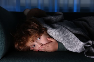 Photo of Afraid boy hiding under blanket on sofa at night