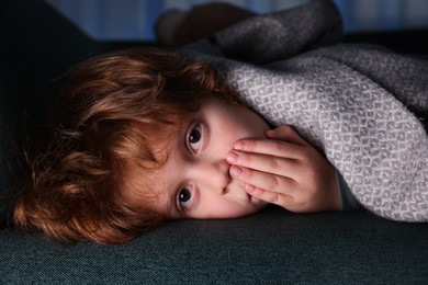 Photo of Afraid boy hiding under blanket on sofa at night