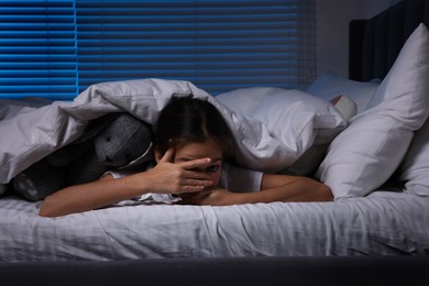 Photo of Fearful girl with toy bunny under duvet in bed at night