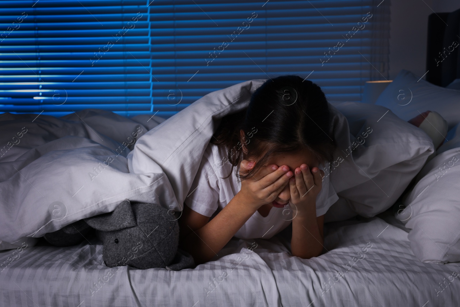 Photo of Fearful girl with toy bunny under duvet in bed at night