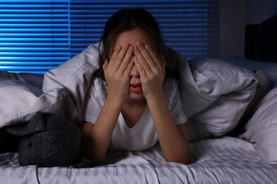 Fearful girl with toy bunny under duvet in bed at night