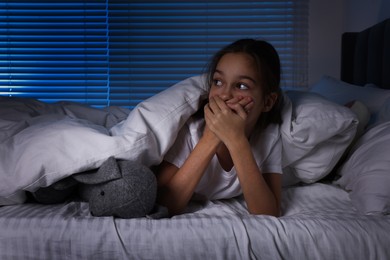 Fearful girl with toy bunny under duvet in bed at night