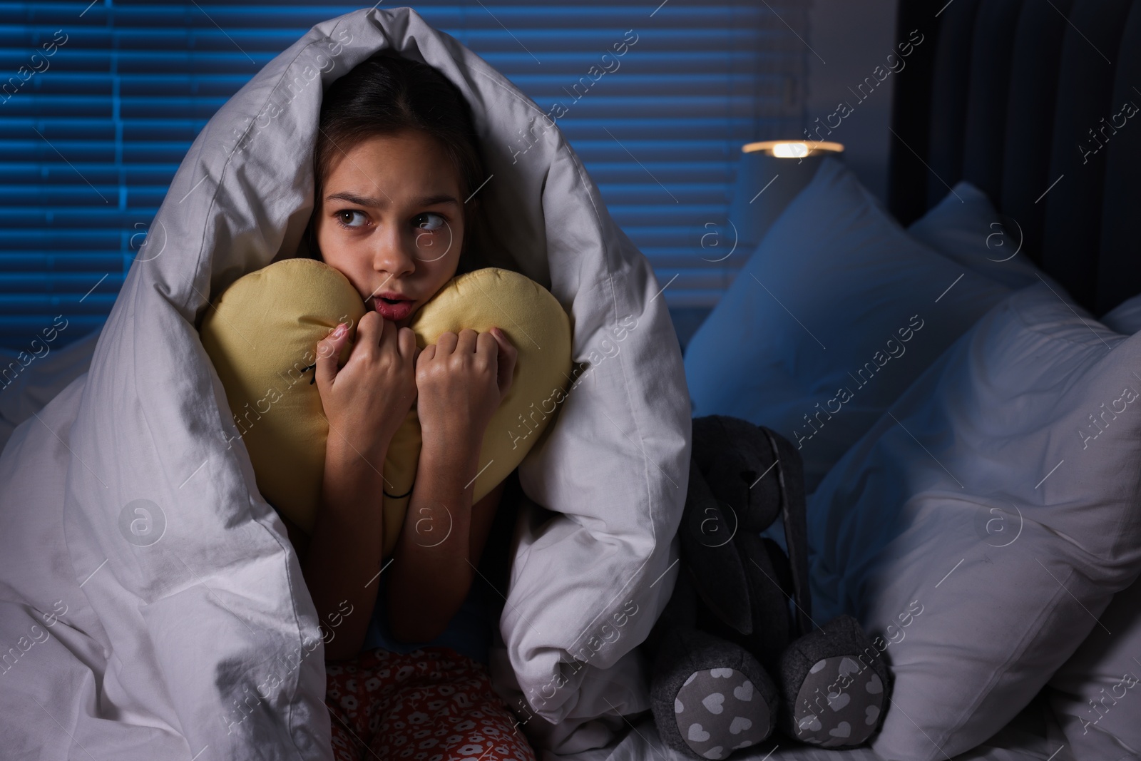 Photo of Afraid girl with pillow under duvet on bed at night