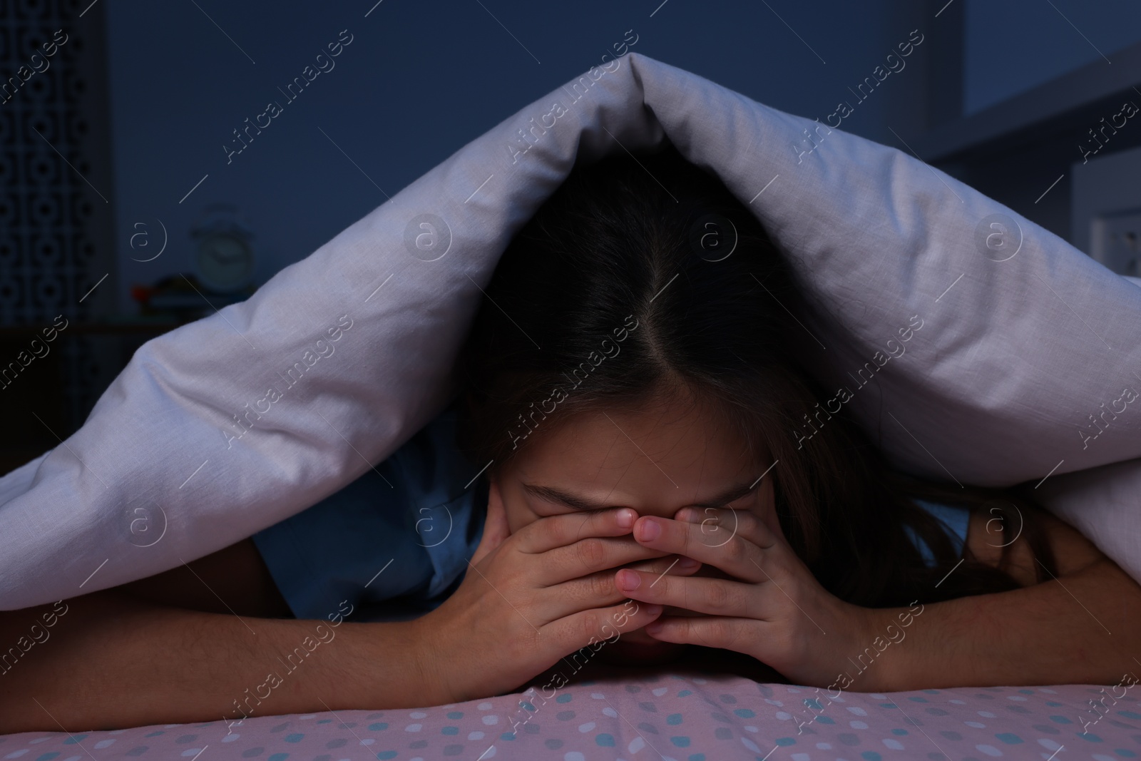 Photo of Afraid girl hiding under duvet on bed at night