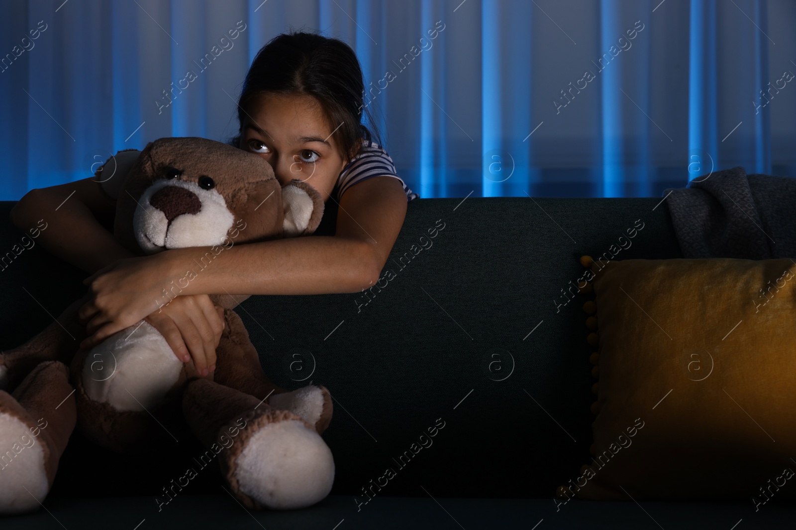 Photo of Scared girl with teddy bear hiding behind sofa at night