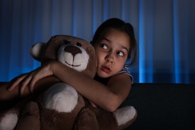 Photo of Scared girl with teddy bear hiding behind sofa at night