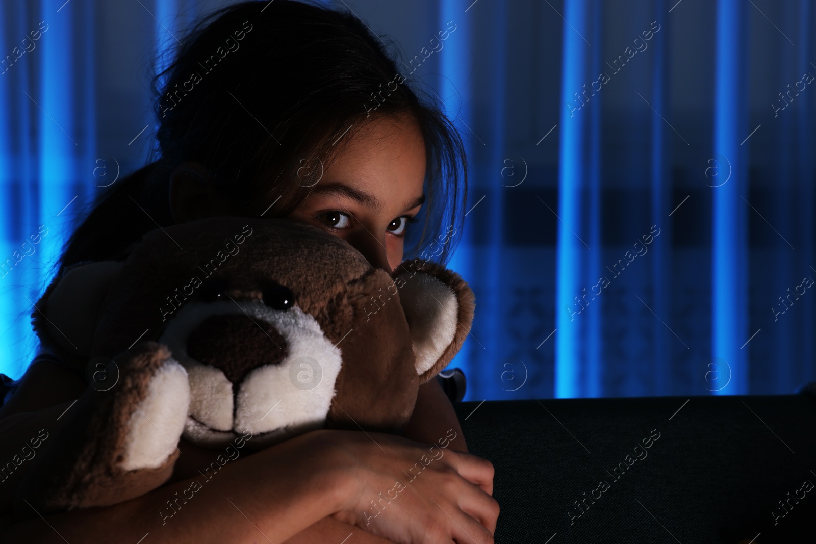 Photo of Scared girl with teddy bear hiding behind sofa at night. Space for text