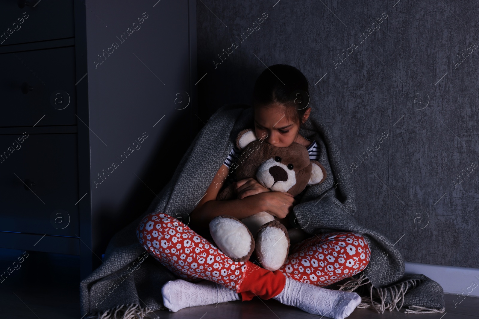 Photo of Scared girl with teddy bear hiding behind chest of drawers in her room at night