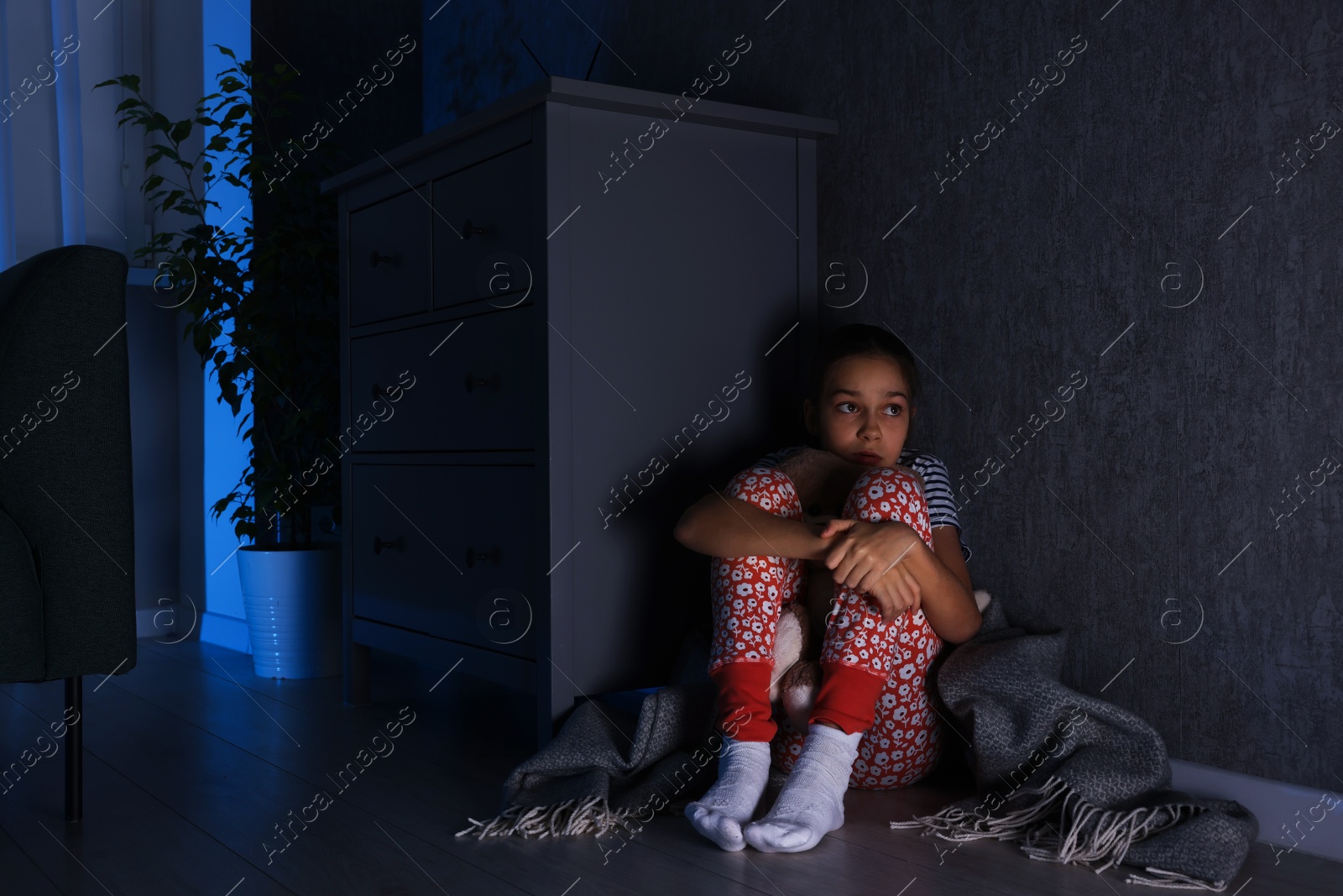 Photo of Scared girl hiding behind chest of drawers in her room at night