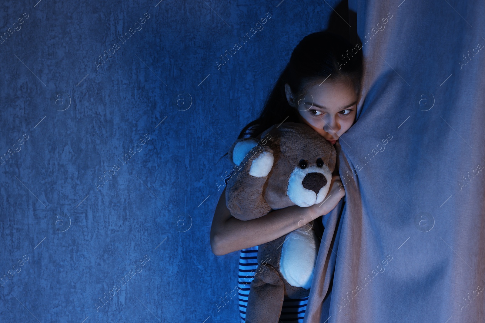 Photo of Scared girl hiding with teddy bear behind curtain at night. Space for text