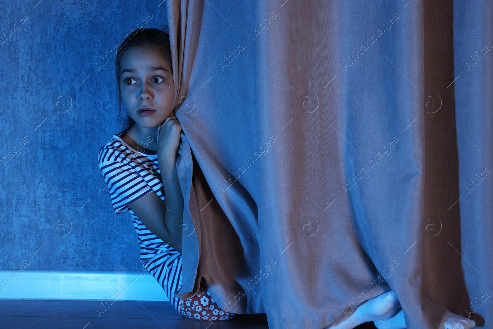 Photo of Scared girl hiding behind curtain at night