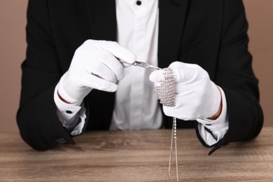 Photo of Appraiser with tweezers evaluating luxury necklace at wooden table, closeup