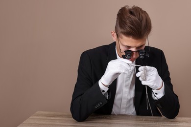 Photo of Appraiser with tweezers evaluating luxury necklace at wooden table against beige background. Space for text