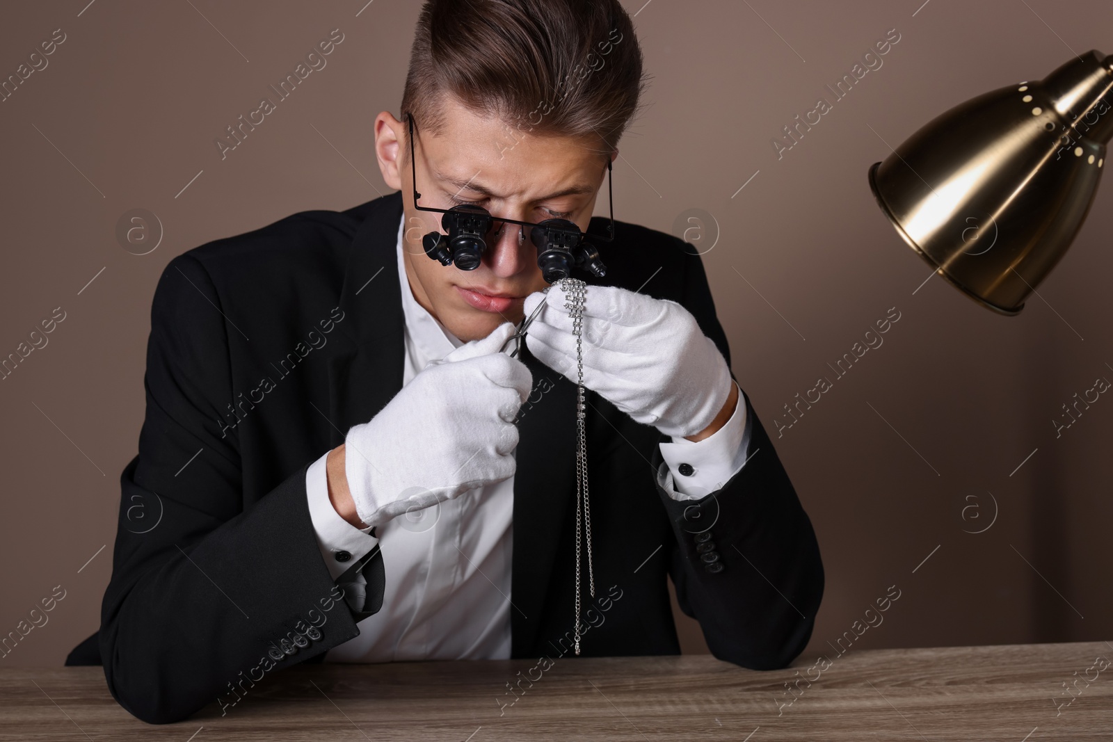 Photo of Appraiser with tweezers evaluating luxury necklace at wooden table against beige background