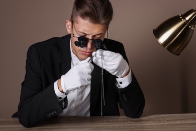 Photo of Appraiser with tweezers evaluating luxury necklace at wooden table against beige background