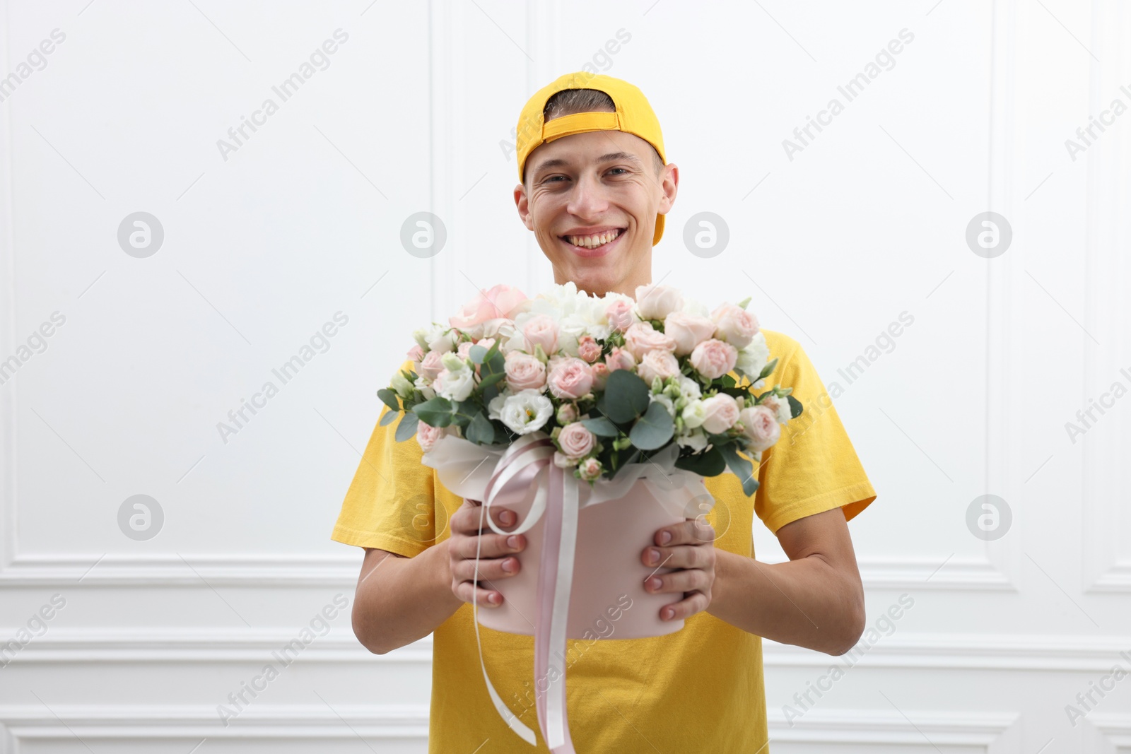 Photo of Smiling delivery man holding gift box with beautiful floral composition near white wall