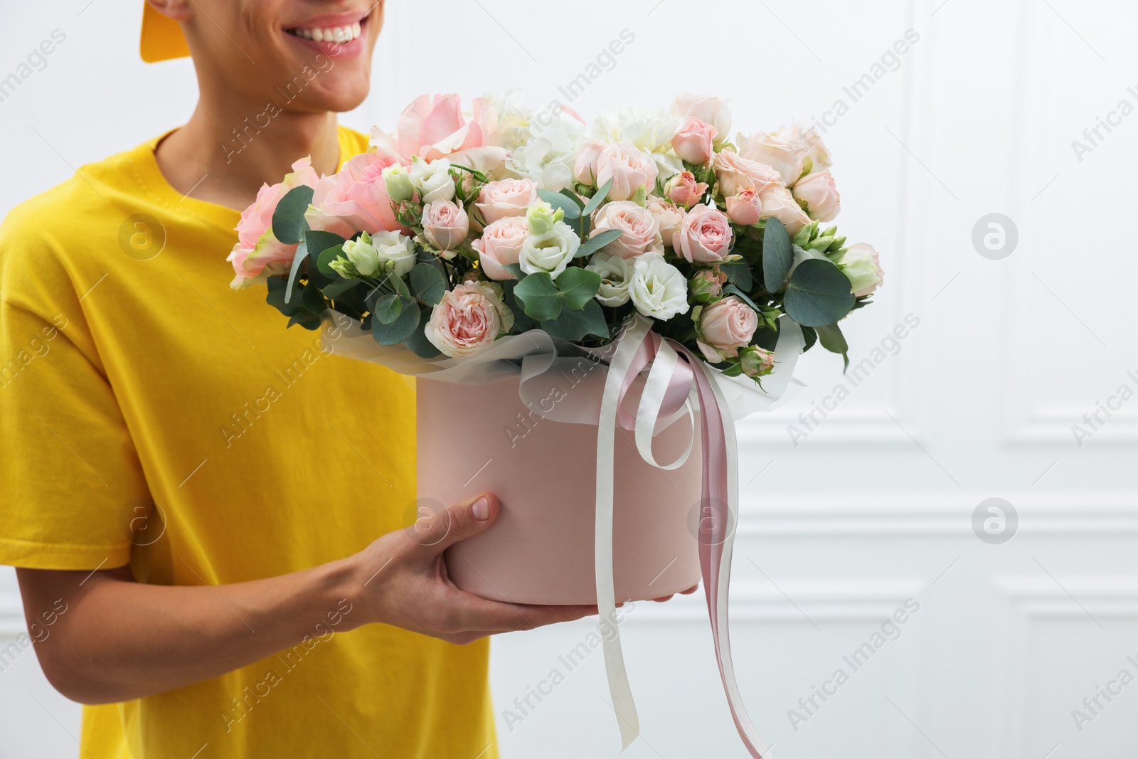 Photo of Smiling delivery man holding gift box with beautiful floral composition near white wall, closeup