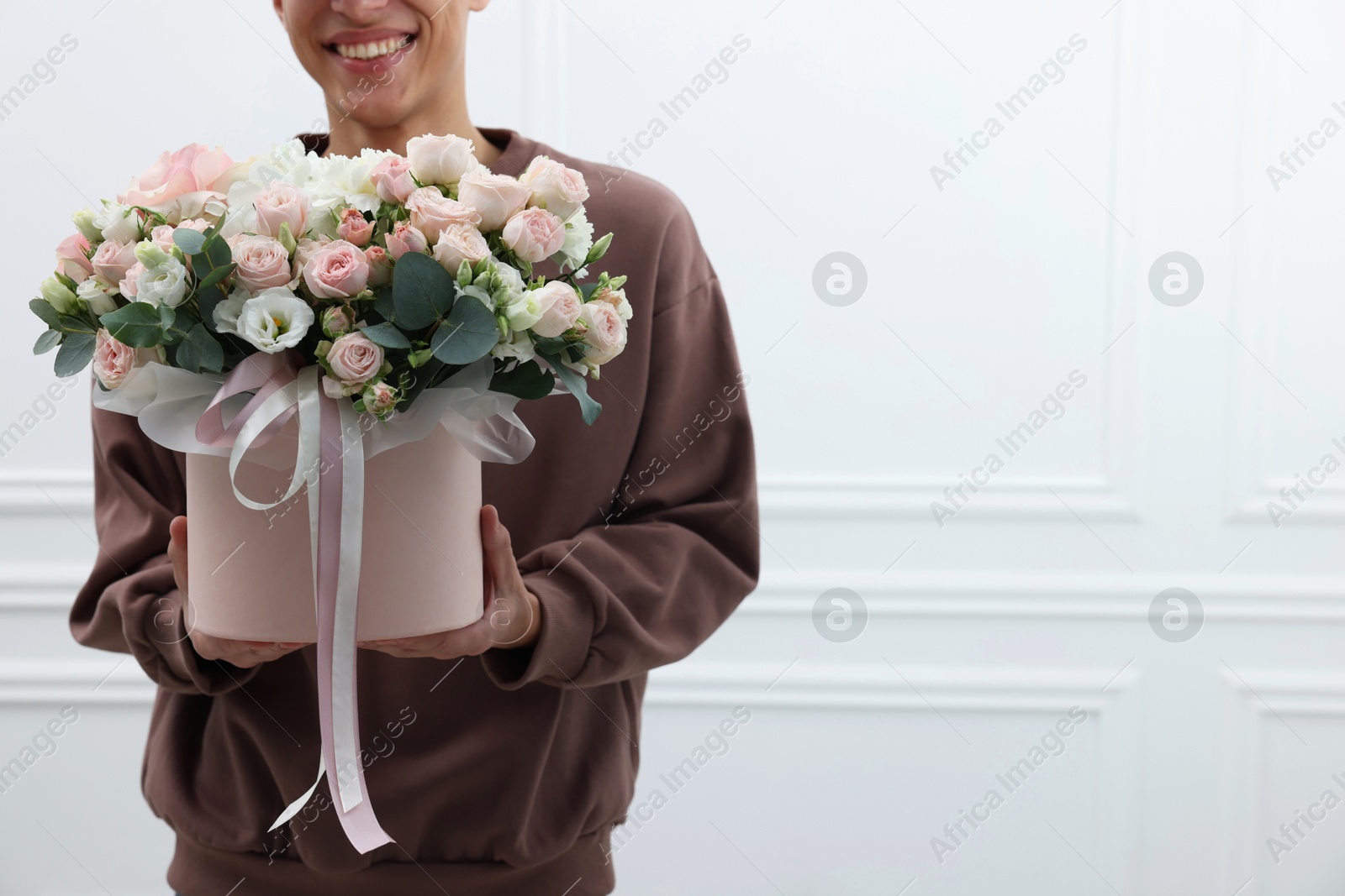 Photo of Smiling delivery man holding gift box with beautiful floral composition near white wall, closeup. Space for text