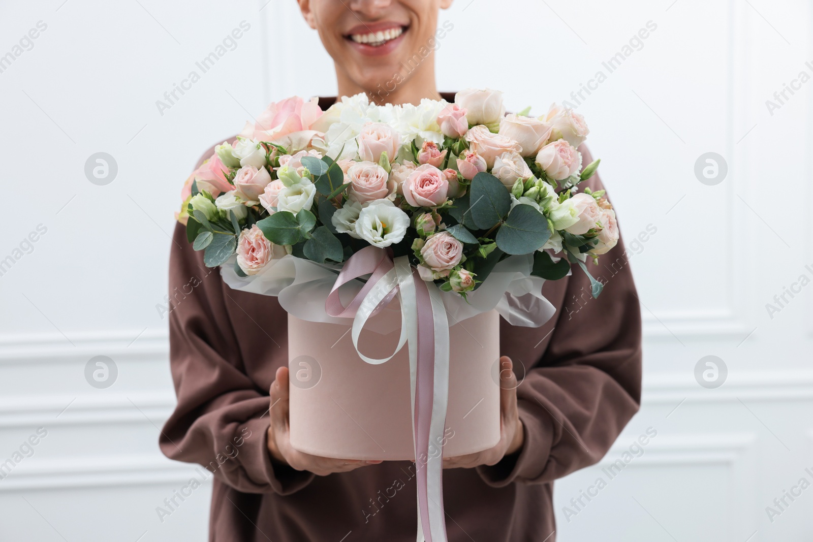 Photo of Smiling delivery man holding gift box with beautiful floral composition near white wall, closeup