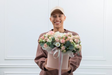 Photo of Smiling delivery man holding gift box with beautiful floral composition near white wall