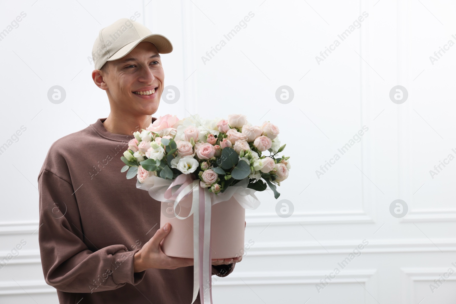 Photo of Smiling delivery man holding gift box with beautiful floral composition near white wall. Space for text