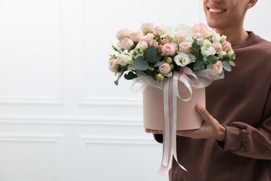 Photo of Smiling delivery man holding gift box with beautiful floral composition near white wall, closeup. Space for text