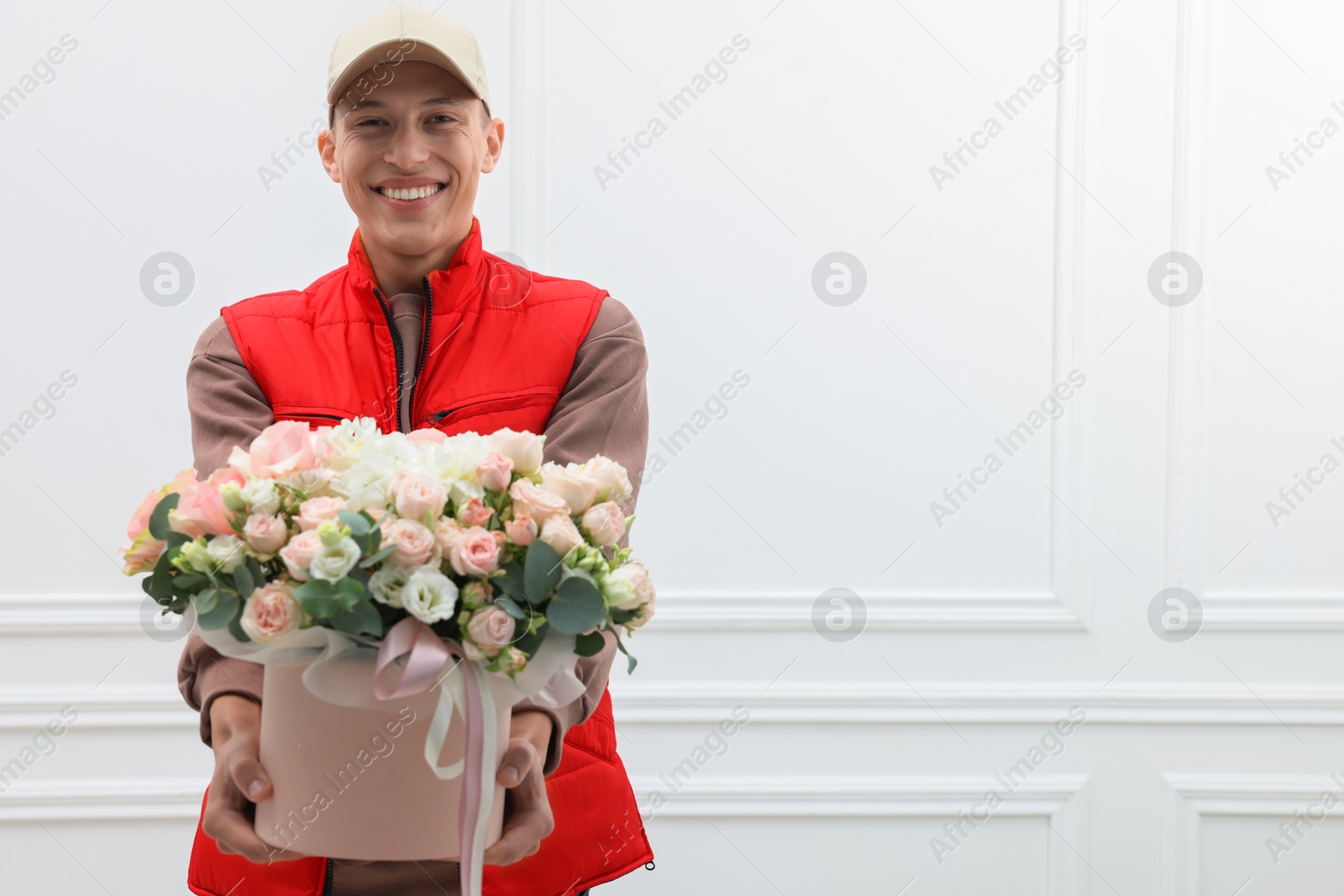 Photo of Smiling delivery man giving gift box with beautiful floral composition near white wall. Space for text