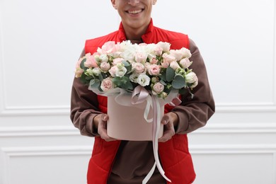 Photo of Smiling delivery man holding gift box with beautiful floral composition near white wall, closeup