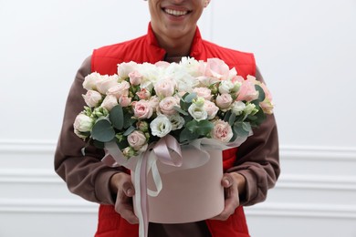 Photo of Smiling delivery man holding gift box with beautiful floral composition near white wall, closeup