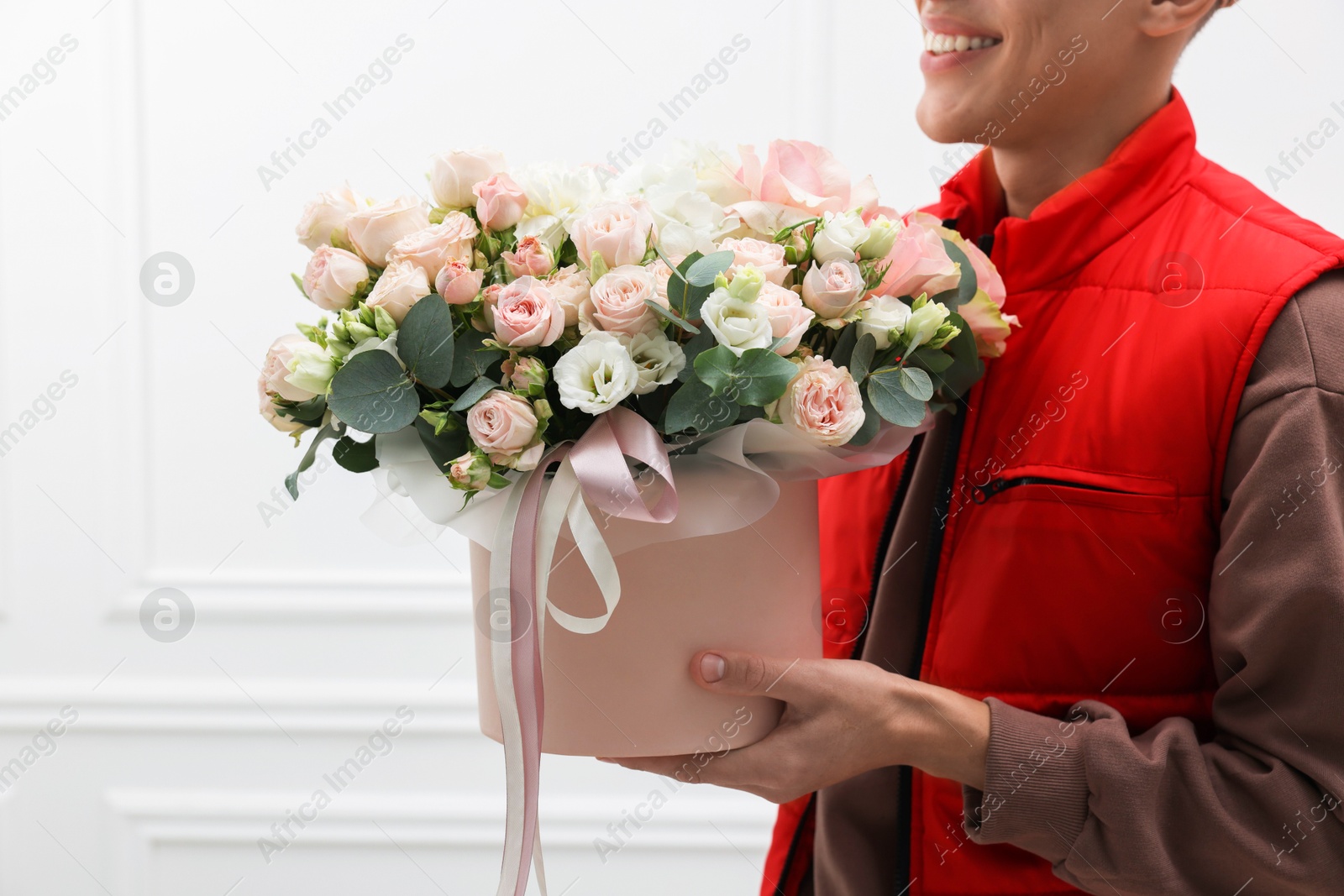 Photo of Smiling delivery man holding gift box with beautiful floral composition near white wall, closeup. Space for text