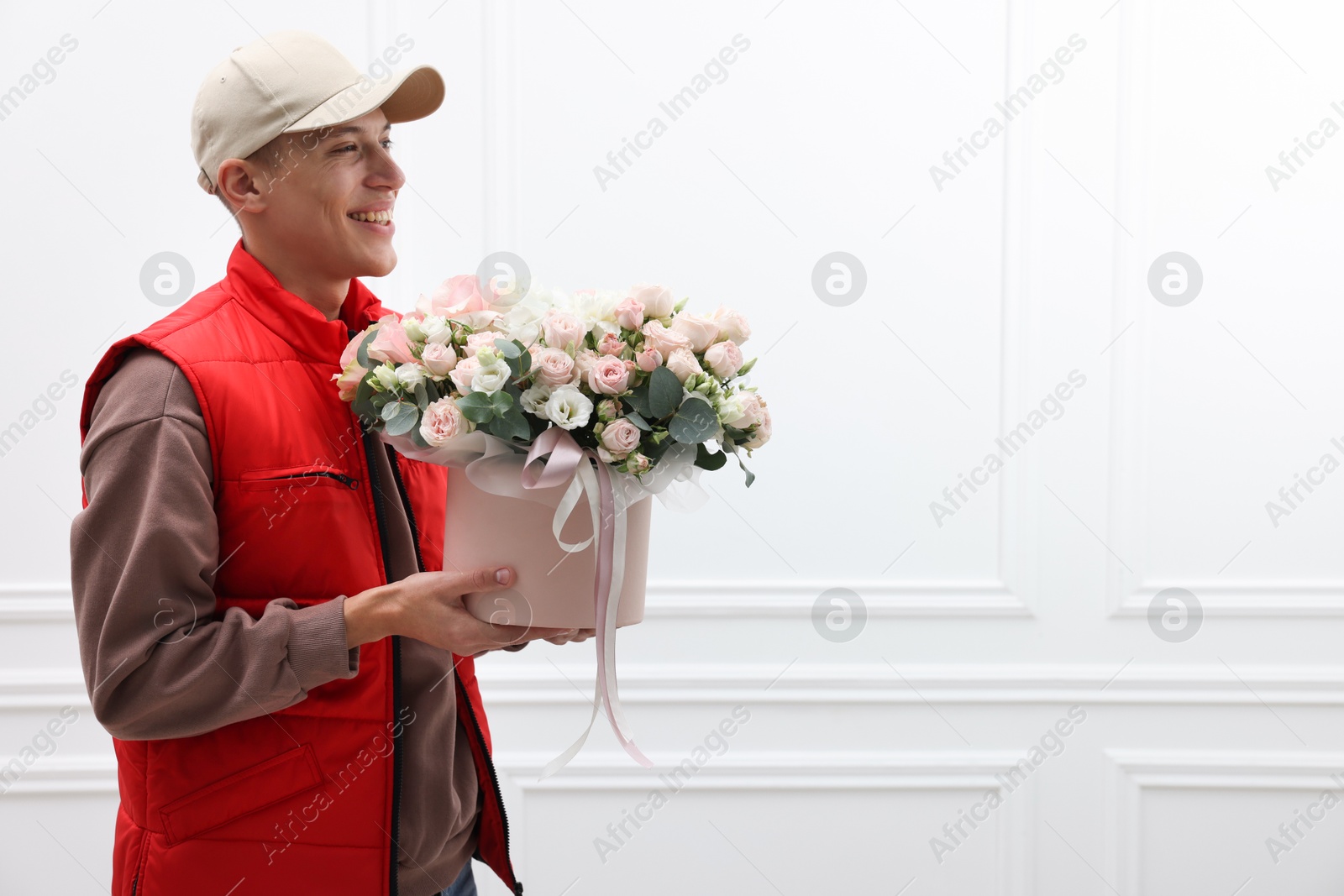 Photo of Smiling delivery man holding gift box with beautiful floral composition near white wall. Space for text