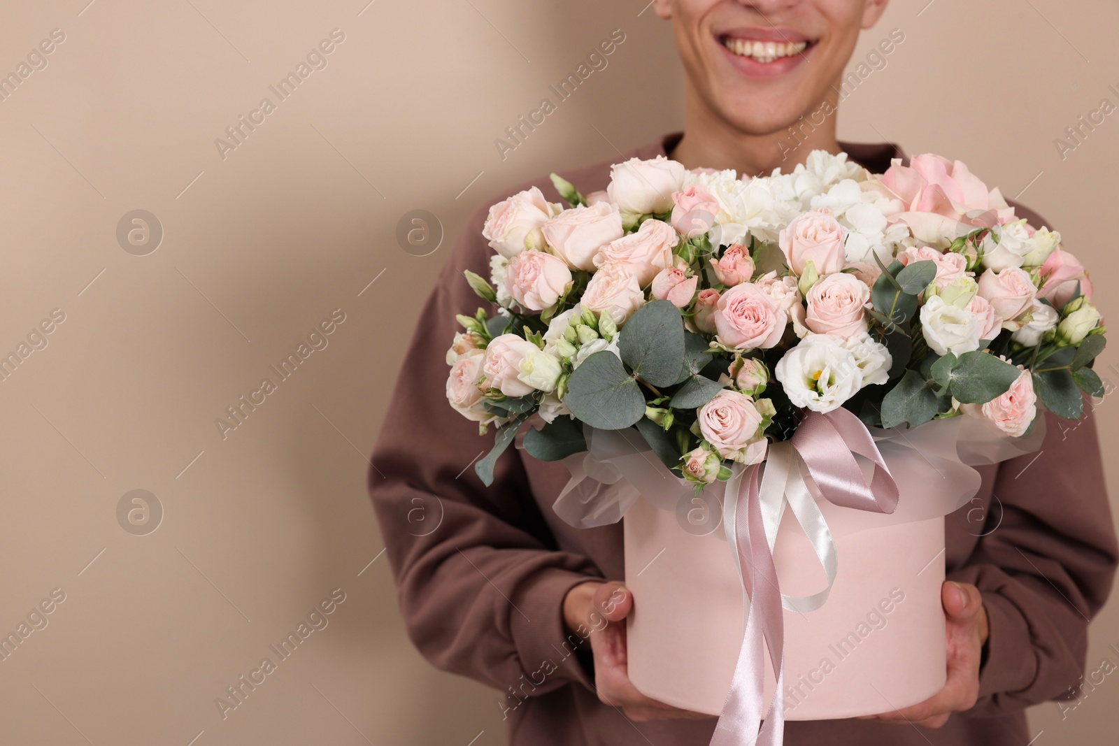 Photo of Smiling delivery man holding gift box with beautiful floral composition on beige background, closeup. Space for text