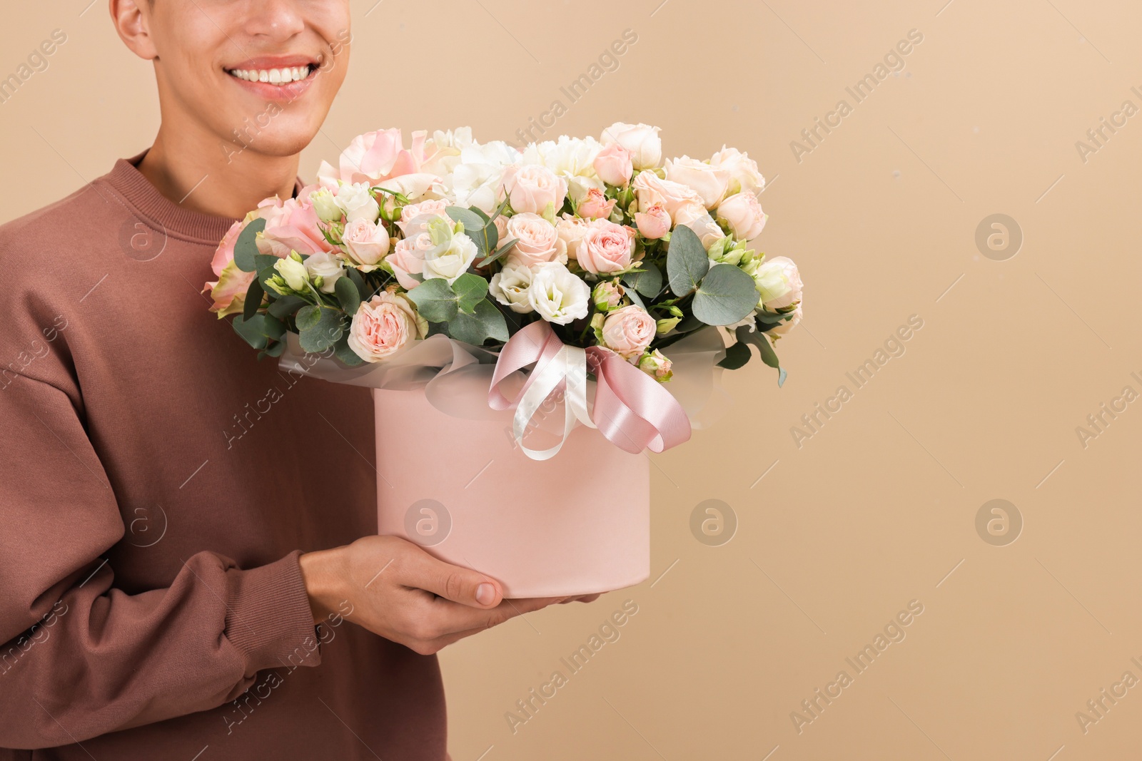 Photo of Smiling delivery man holding gift box with beautiful floral composition on beige background, closeup. Space for text