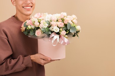 Smiling delivery man holding gift box with beautiful floral composition on beige background, closeup. Space for text