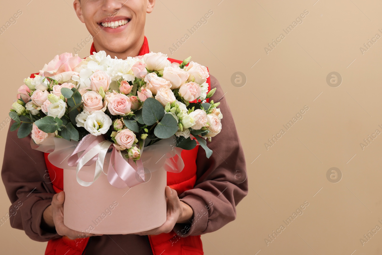 Photo of Smiling delivery man holding gift box with beautiful floral composition on beige background, closeup. Space for text