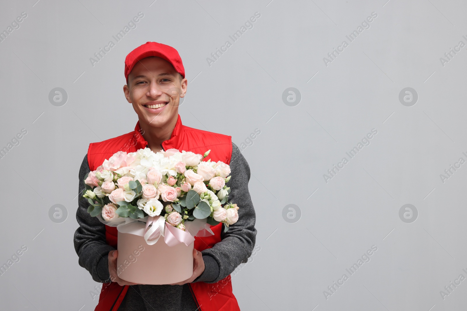 Photo of Smiling delivery man holding gift box with beautiful floral composition on grey background. Space for text