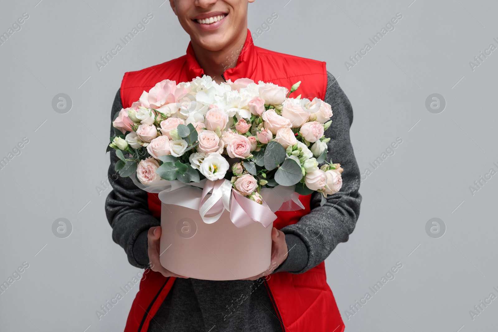 Photo of Smiling delivery man holding gift box with beautiful floral composition on grey background, closeup