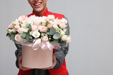 Photo of Smiling delivery man holding gift box with beautiful floral composition on grey background, closeup. Space for text