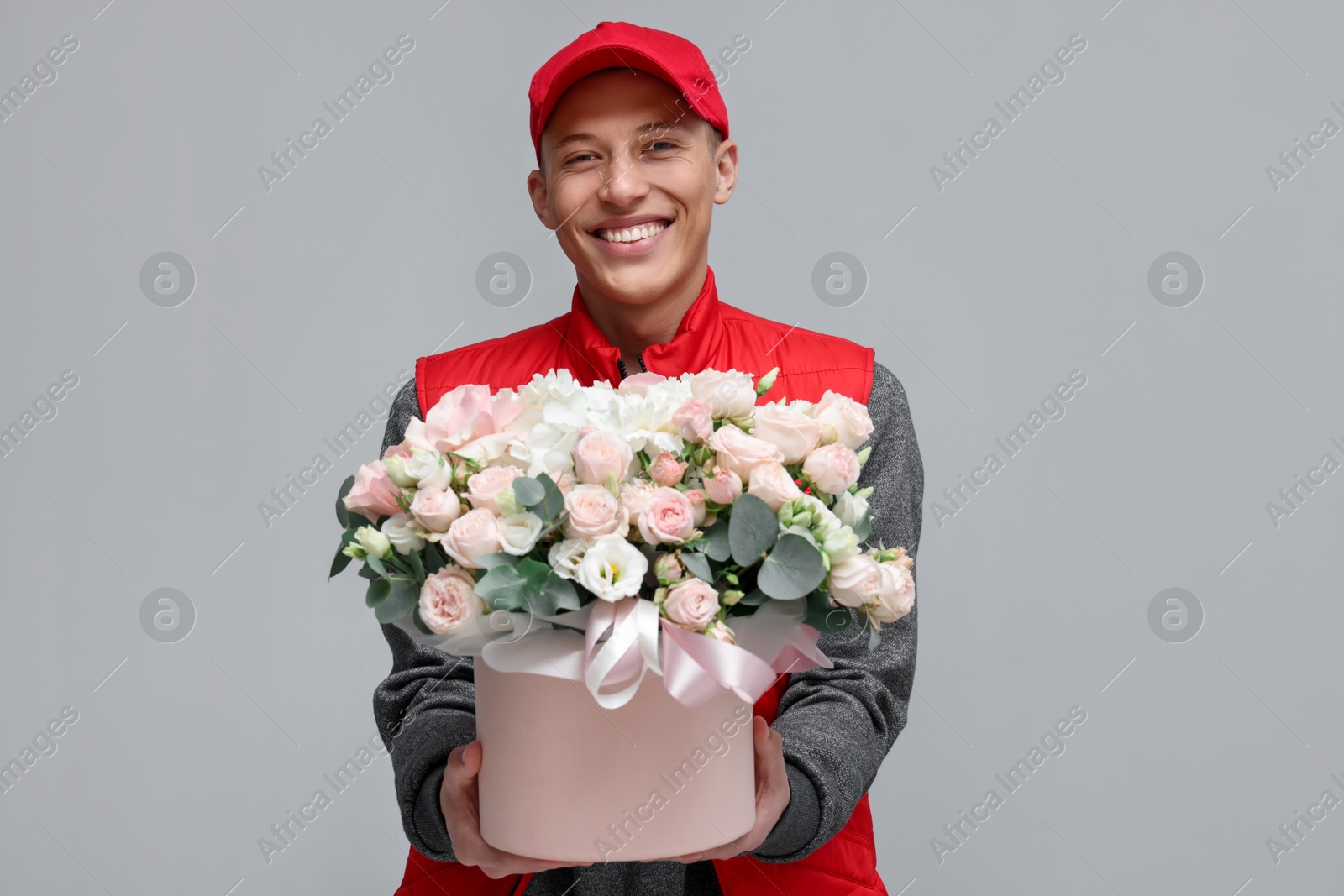 Photo of Smiling delivery man holding gift box with beautiful floral composition on grey background