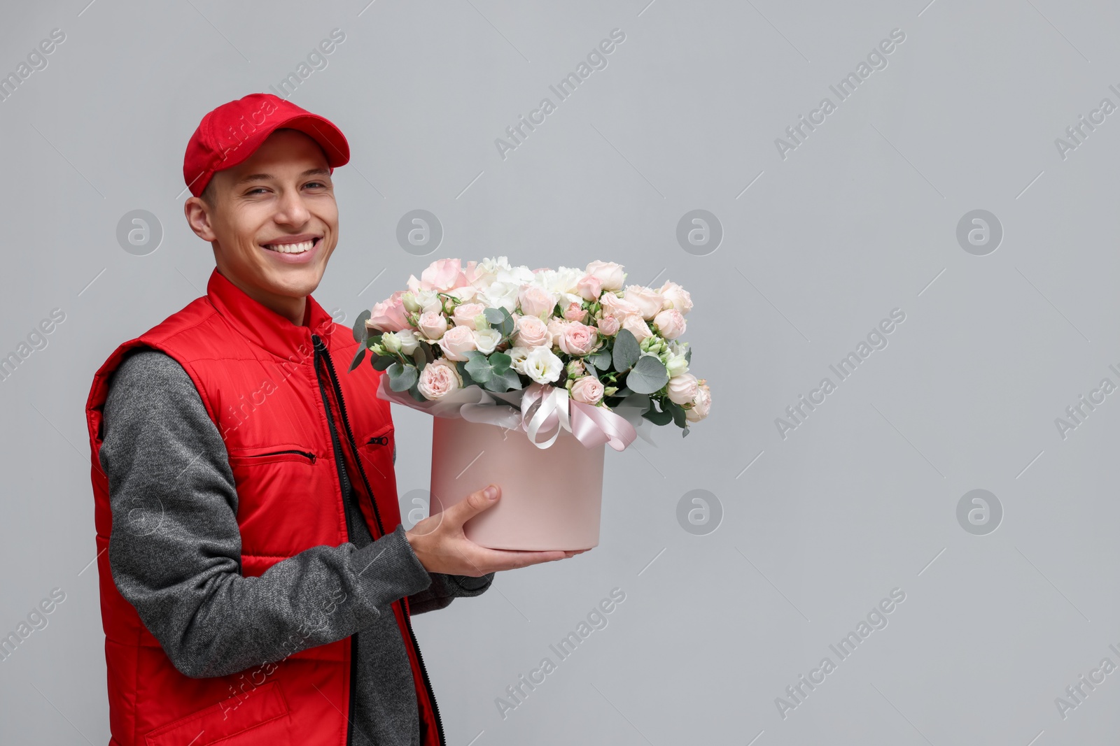 Photo of Smiling delivery man holding gift box with beautiful floral composition on grey background. Space for text