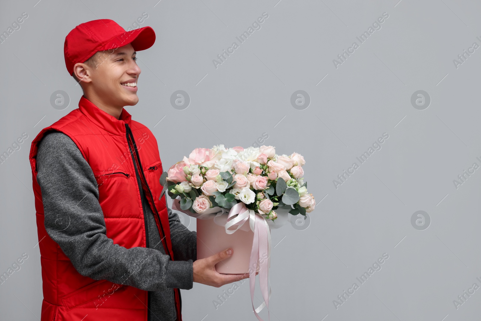 Photo of Smiling delivery man holding gift box with beautiful floral composition on grey background. Space for text