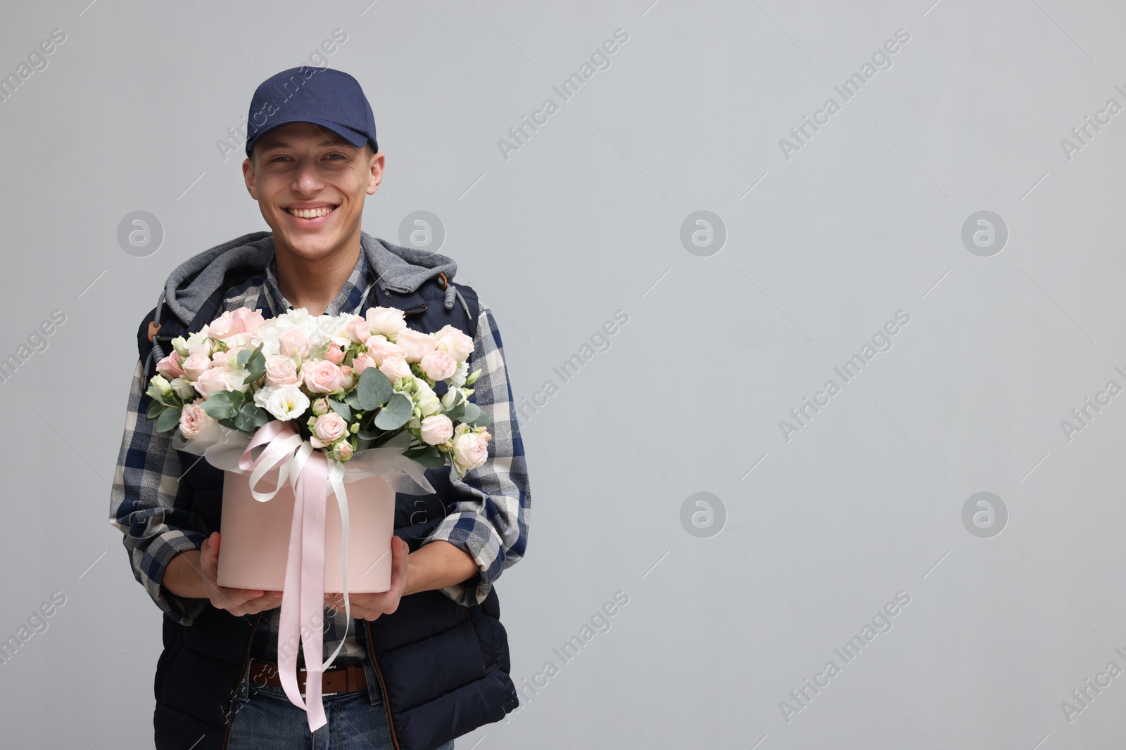 Photo of Smiling delivery man holding gift box with beautiful floral composition on grey background. Space for text