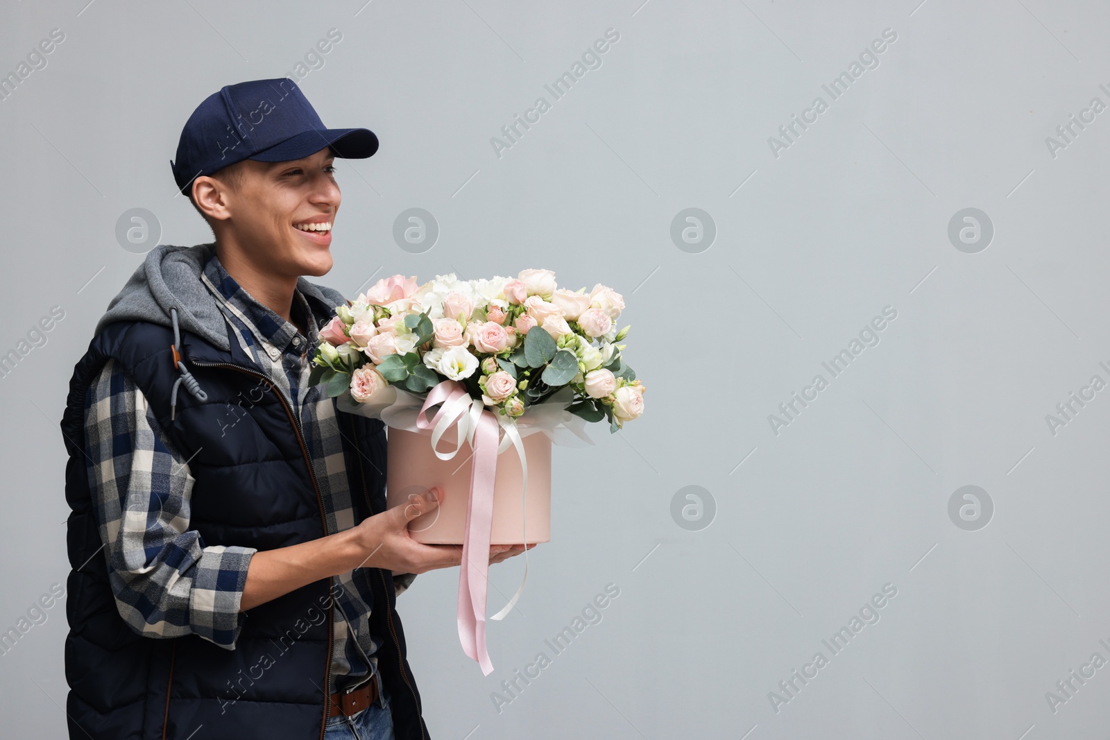 Photo of Smiling delivery man holding gift box with beautiful floral composition on grey background. Space for text